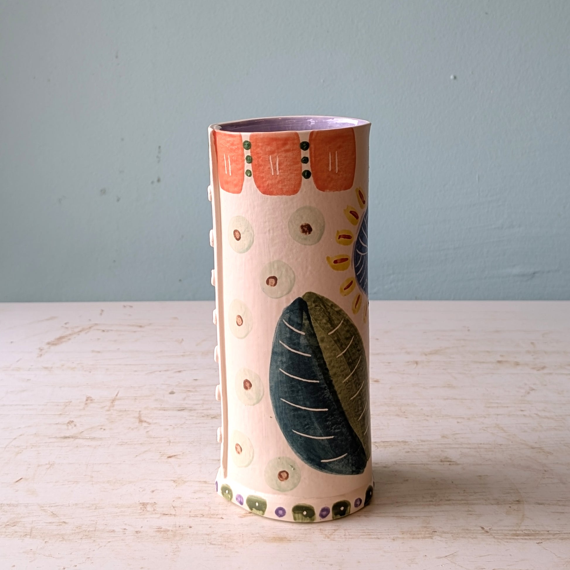 Cylindrical ceramic vase with floral and leaf patterns on a light wooden surface and blue background