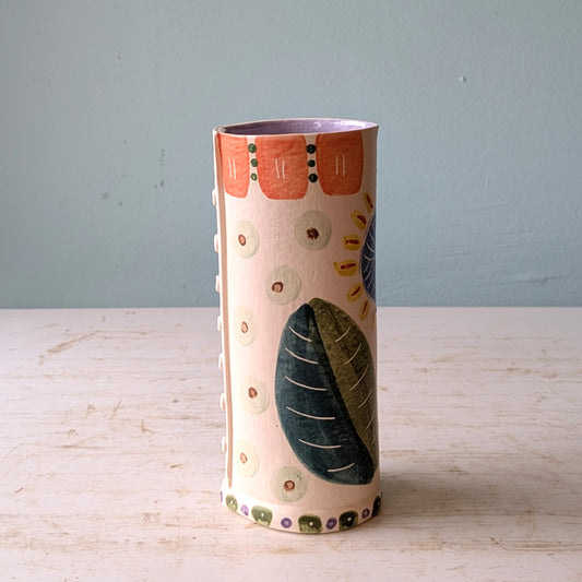Cylindrical ceramic vase with floral and leaf patterns on a light wooden surface and blue background