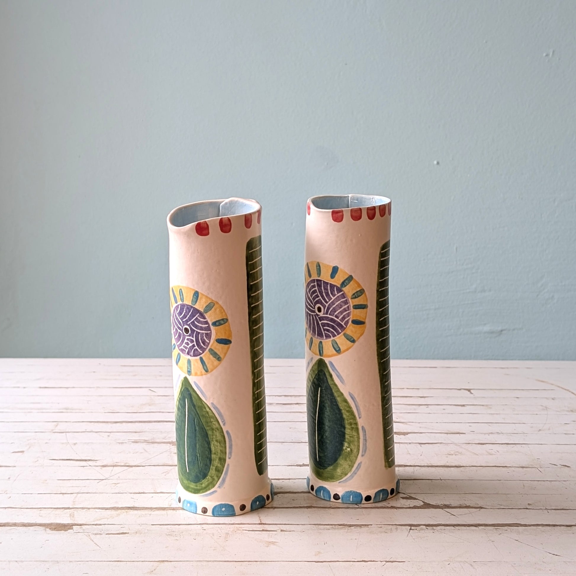Front view of two colourful ceramic pourers on a white wooden surface against a blue background.