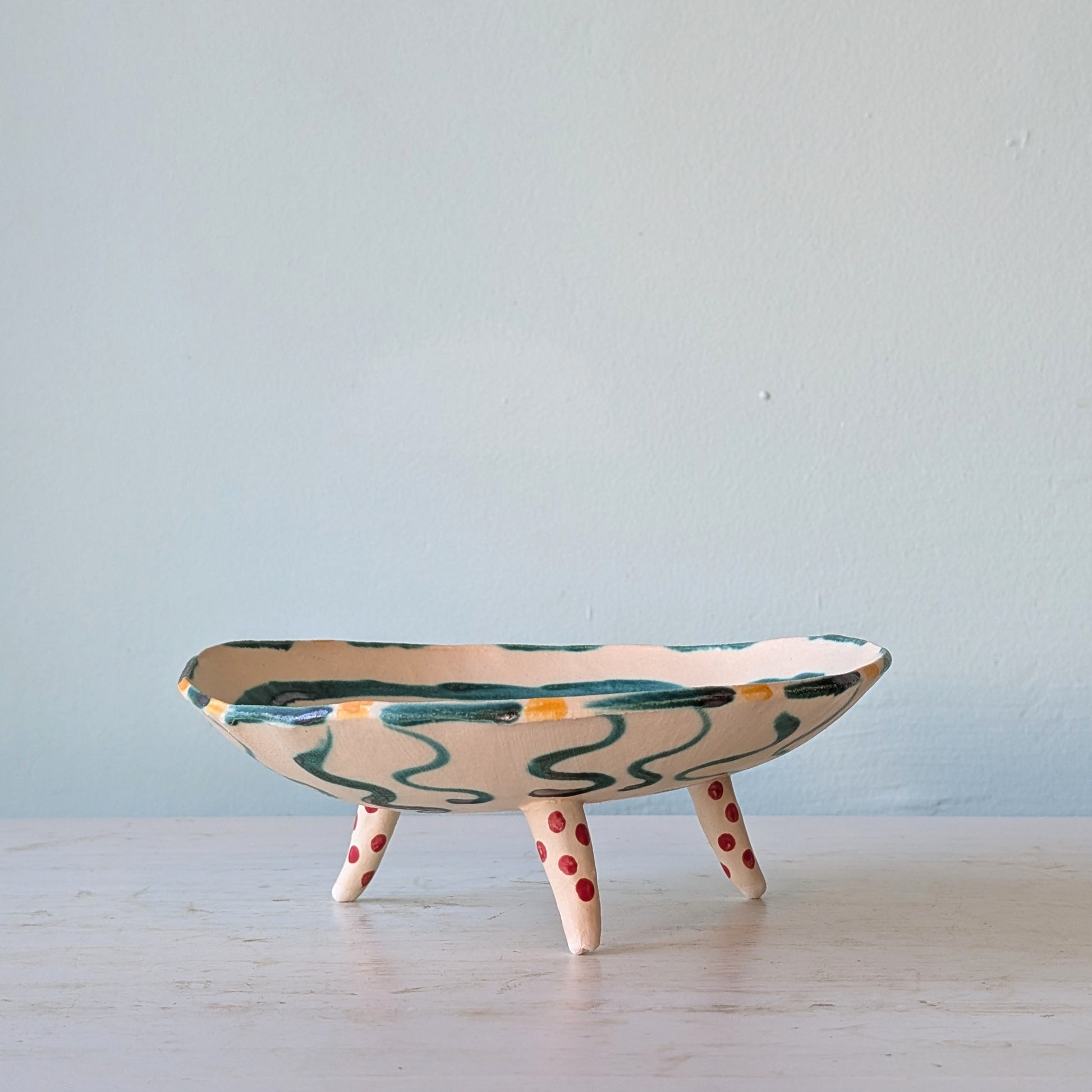 A handmade ceramic shallow bowl with three legs, featuring a cream-colored glaze with green wavy patterns and red polka dots on the legs, set against a light blue wall.