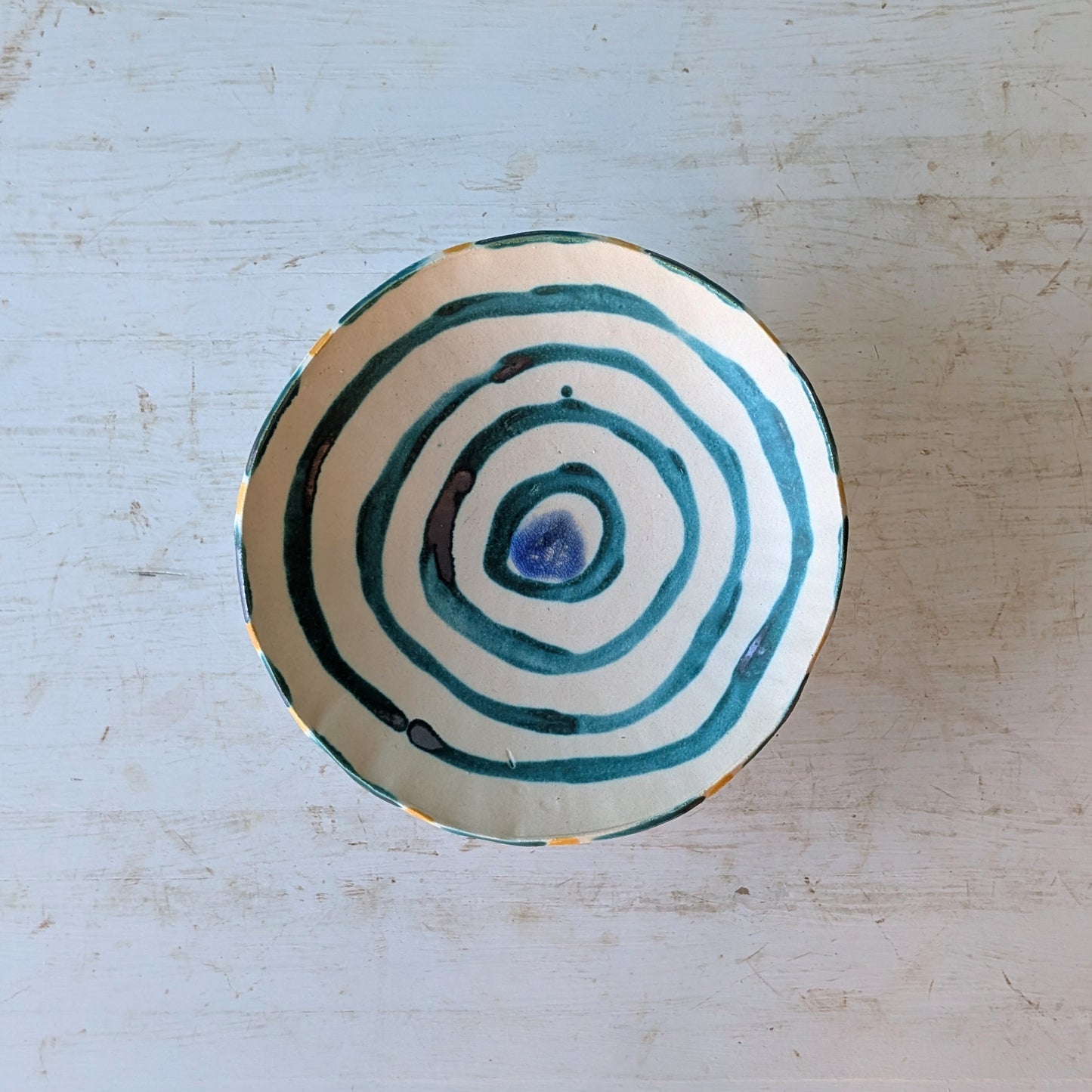 A handmade ceramic bowl with a hand-painted teal and blue spiral pattern on a cream-colored glaze, photographed from directly above on a distressed white wooden surface.