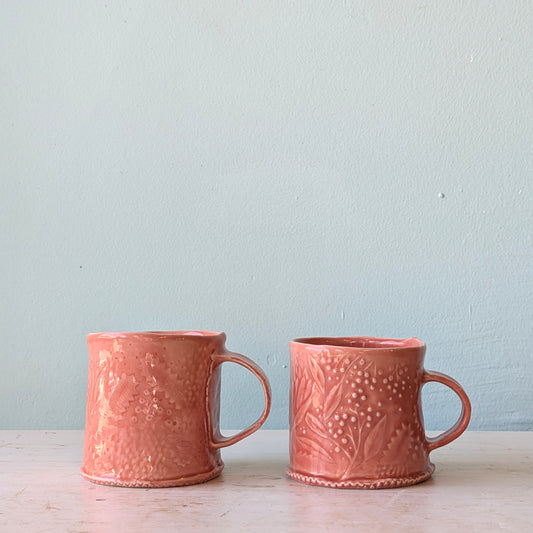Two coral pink ceramic mugs with embossed floral patterns, sitting side-by-side on a light-colored surface against a pale blue wall.