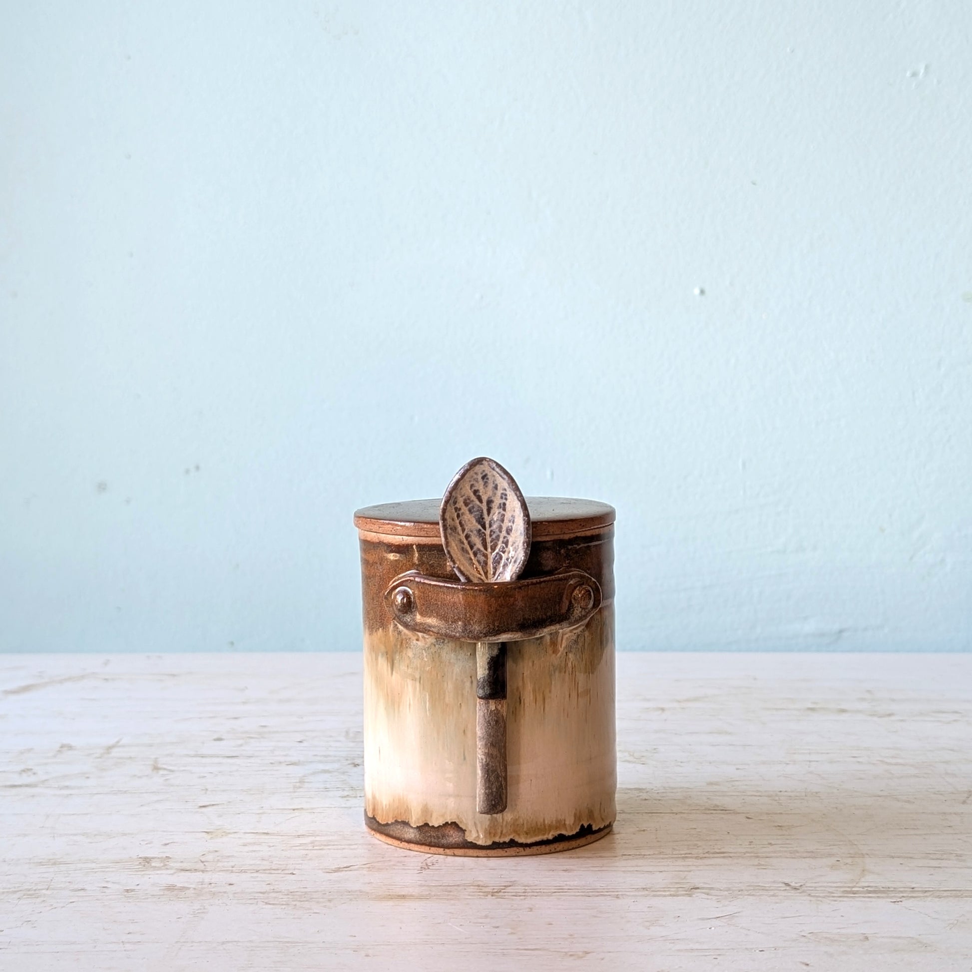 Ceramic canister with a lid and attached spoon on a light wooden surface with a light blue background