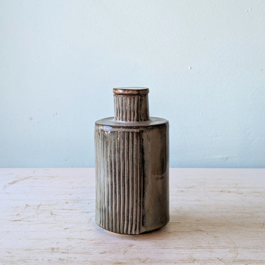 Cylindrical ceramic bottle with ribbed texture and teardrop shaped lid on a light wooden surface and light blue background