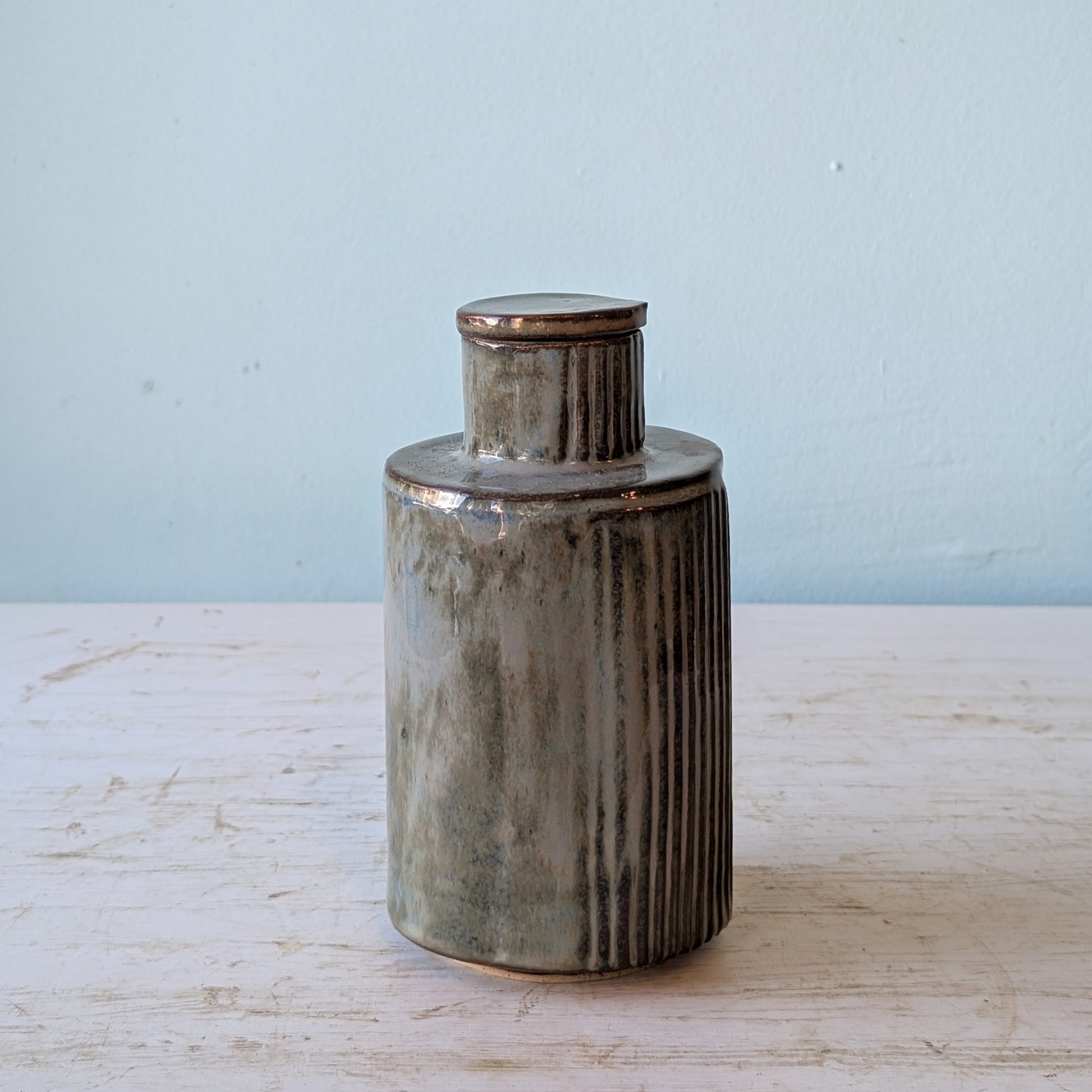 Cylindrical ceramic bottle with ribbed texture and teardrop shaped stopper on a light wooden surface and blue background
