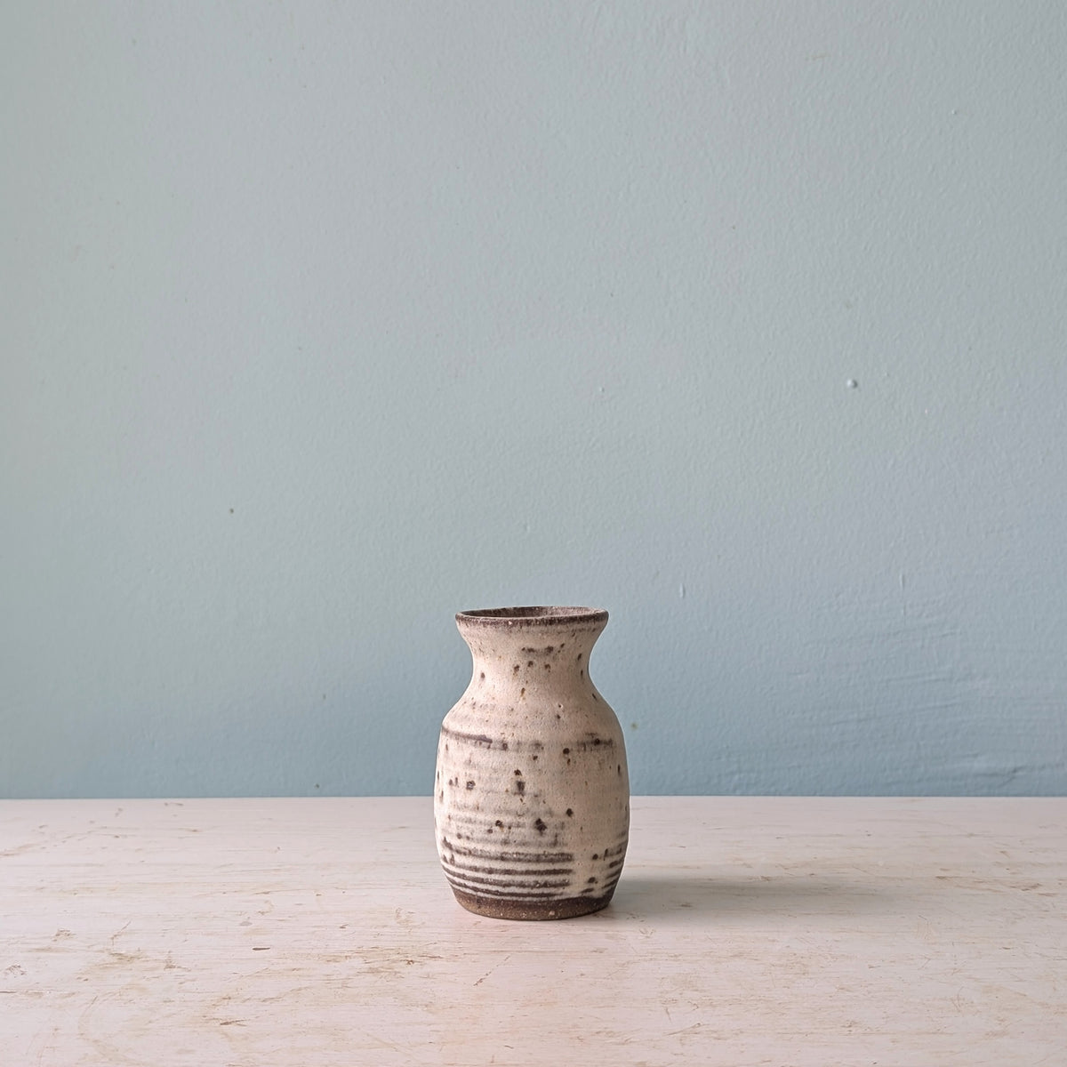 A small handmade ceramic vase with a textured, speckled beige and brown glaze, standing on a light wooden surface against a muted blue wall.