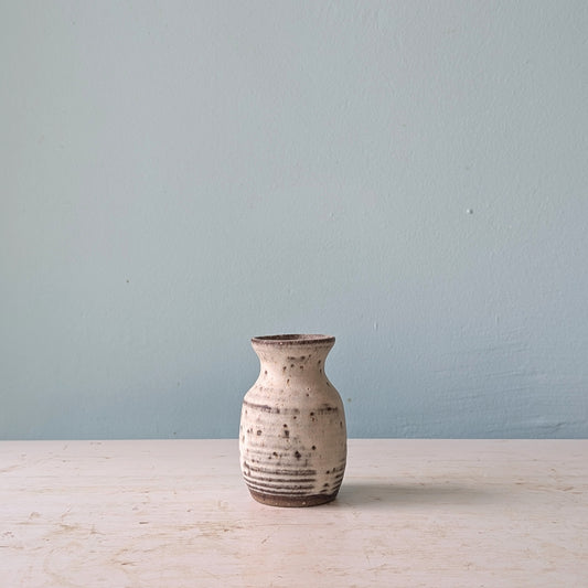 A small handmade ceramic vase with a textured, speckled beige and brown glaze, standing on a light wooden surface against a muted blue wall.