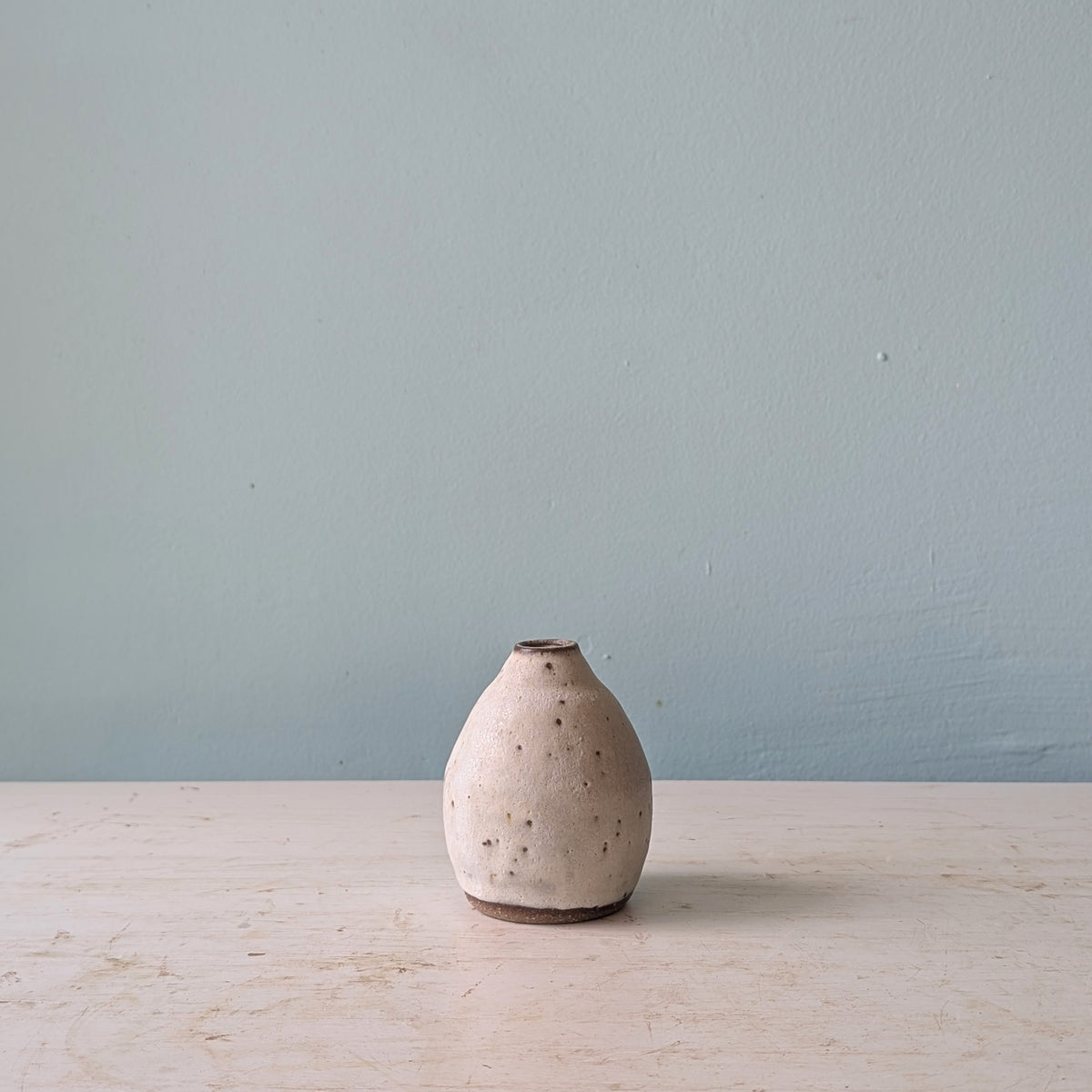 A small handmade ceramic vase with a speckled beige and white glaze, standing on a light wooden surface against a muted blue wall.