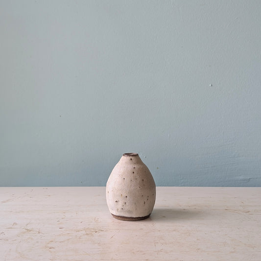 A small handmade ceramic vase with a speckled beige and white glaze, standing on a light wooden surface against a muted blue wall.