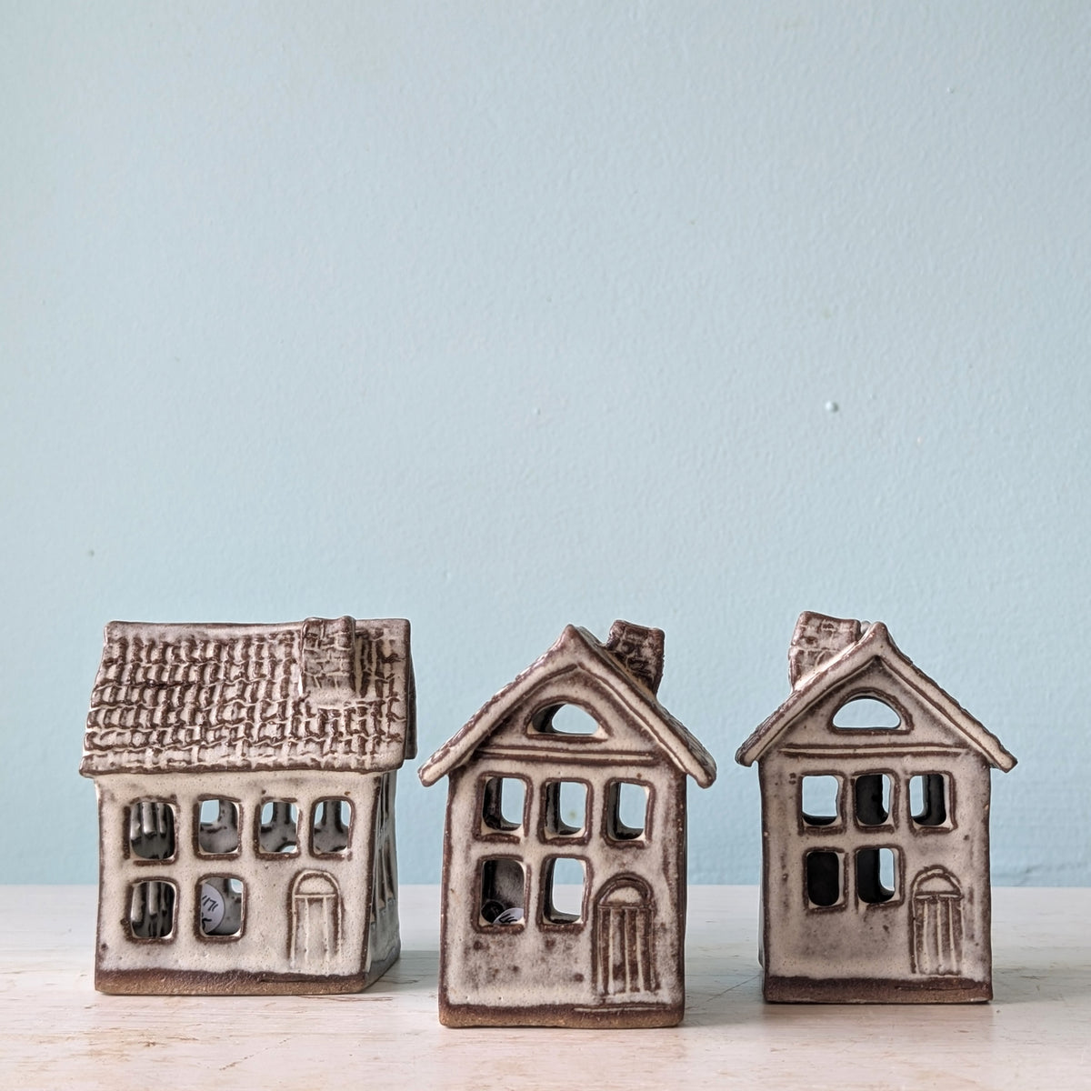 Three small decorative house figurines with textured stoneware-like surfaces in warm beige and brown tones, arranged on a light surface against a pale blue wall.