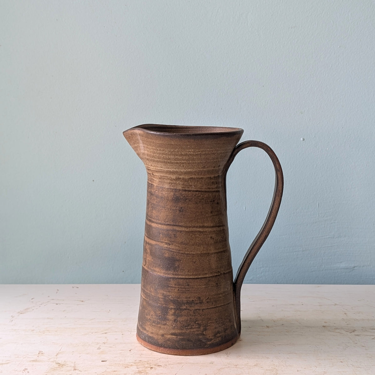 A handmade ceramic pitcher with a rustic, ribbed brown glaze, featuring a wide mouth and a curved handle, set against a light blue wall and a light wood surface.