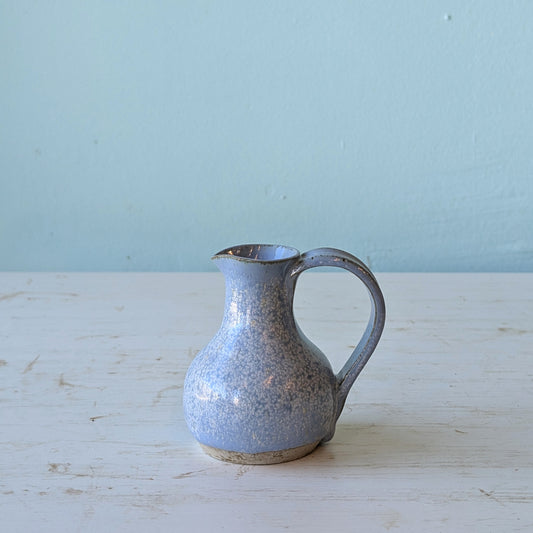 Speckled blue ceramic pitcher on a light wooden surface with a light blue background