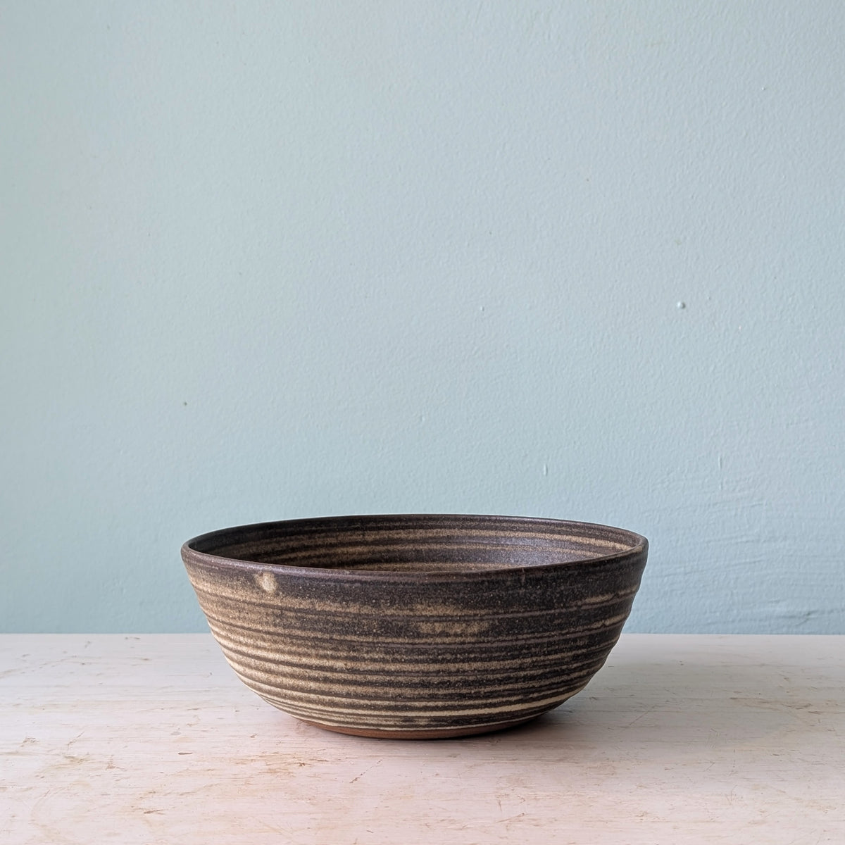 A handmade dark brown ceramic bowl on a white wooden table against a blue wall.