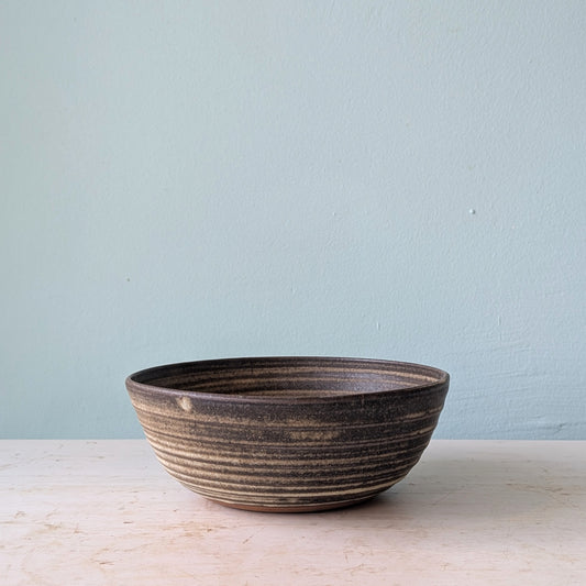 A handmade dark brown ceramic bowl on a white wooden table against a blue wall.