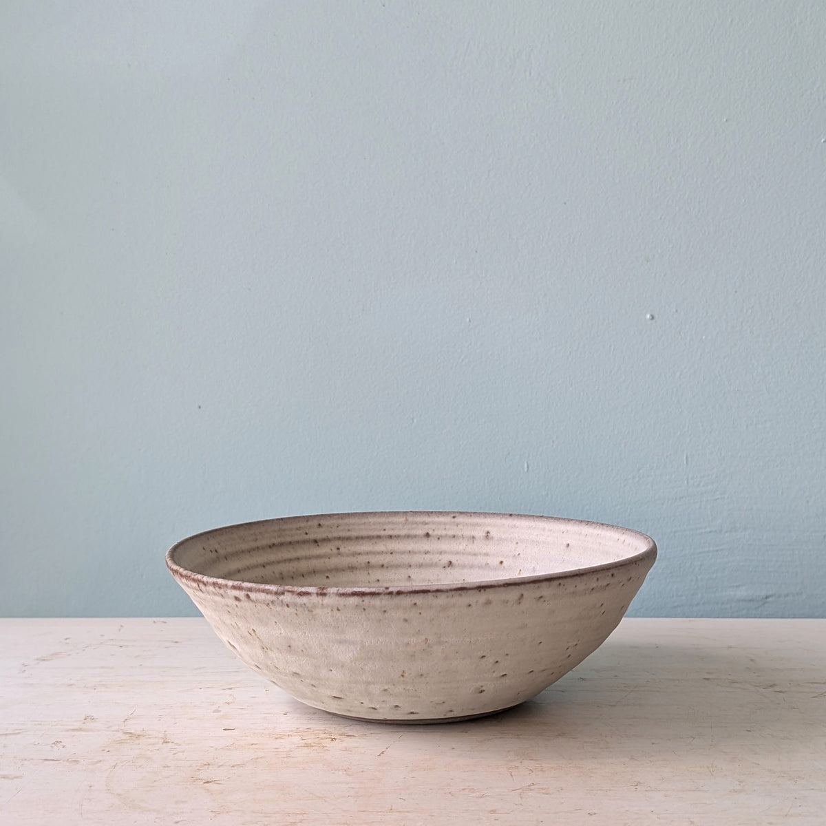 A handmade ceramic bowl with a textured, speckled off-white glaze and a rustic brown rim, sitting on a light wooden surface against a pale blue wall.