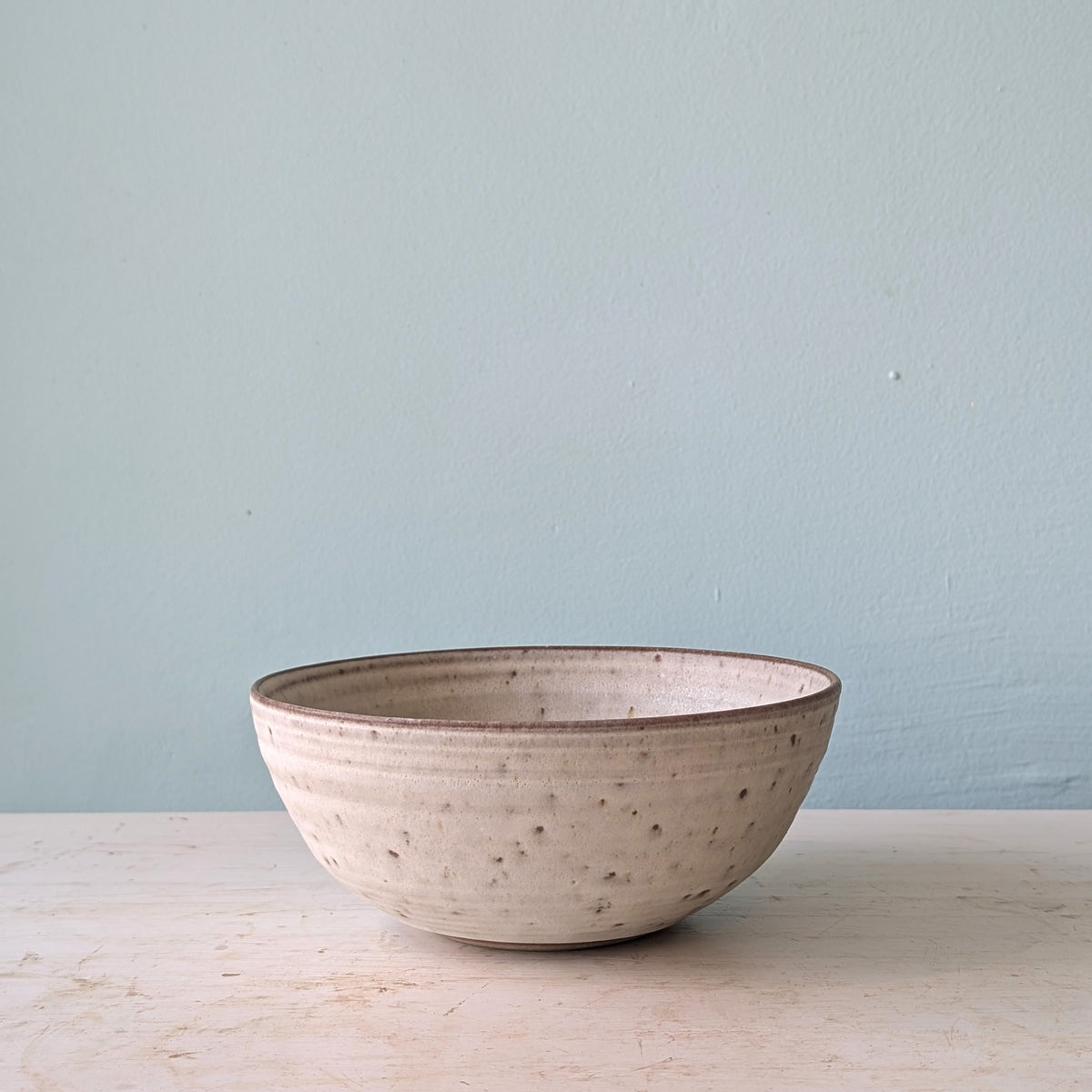 A handmade ceramic bowl with a speckled, textured off-white glaze and a dark brown rim, sitting on a light wooden surface against a pale blue wall.