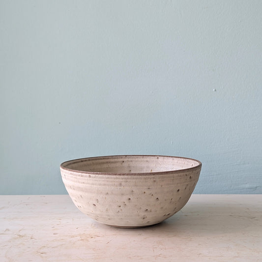 A handmade ceramic bowl with a speckled, textured off-white glaze and a dark brown rim, sitting on a light wooden surface against a pale blue wall.