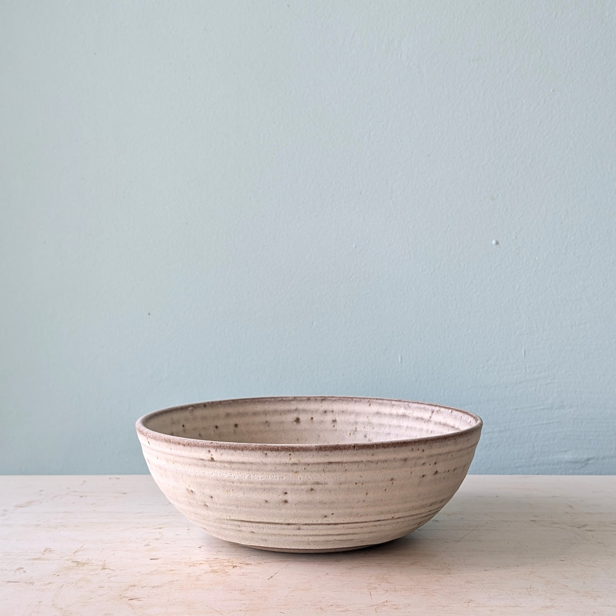 A handmade ceramic bowl with a speckled beige and brown glaze, sitting on a light wooden surface against a pale blue wall.