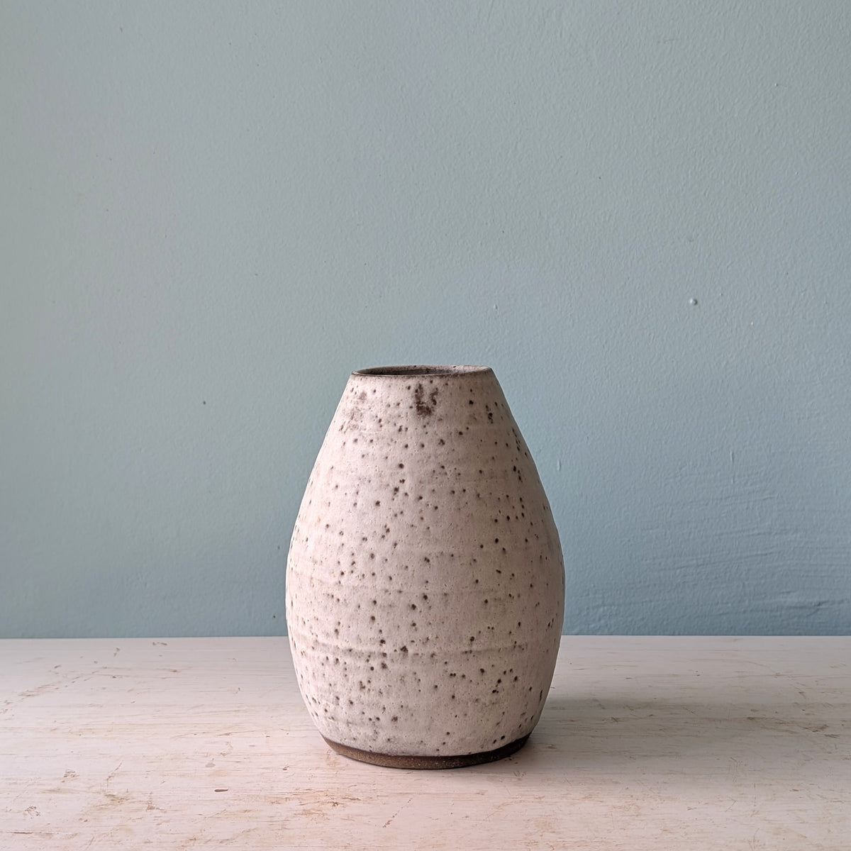 A small handmade ceramic vase with a textured, speckled off-white glaze, standing on a light wooden surface against a muted blue wall.