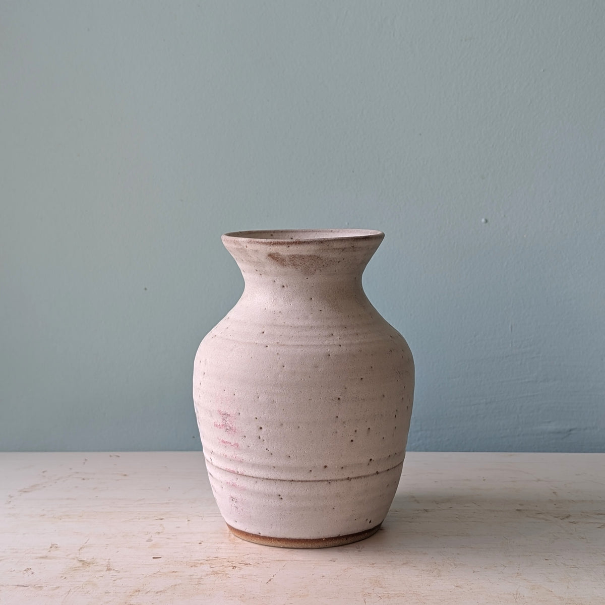 A handmade ceramic vase with a textured, speckled off-white glaze, standing on a light wooden surface against a muted blue wall.