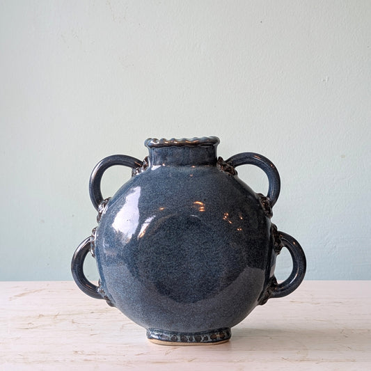 Blue ceramic vase with four looped handles on a white table against a blue wall.