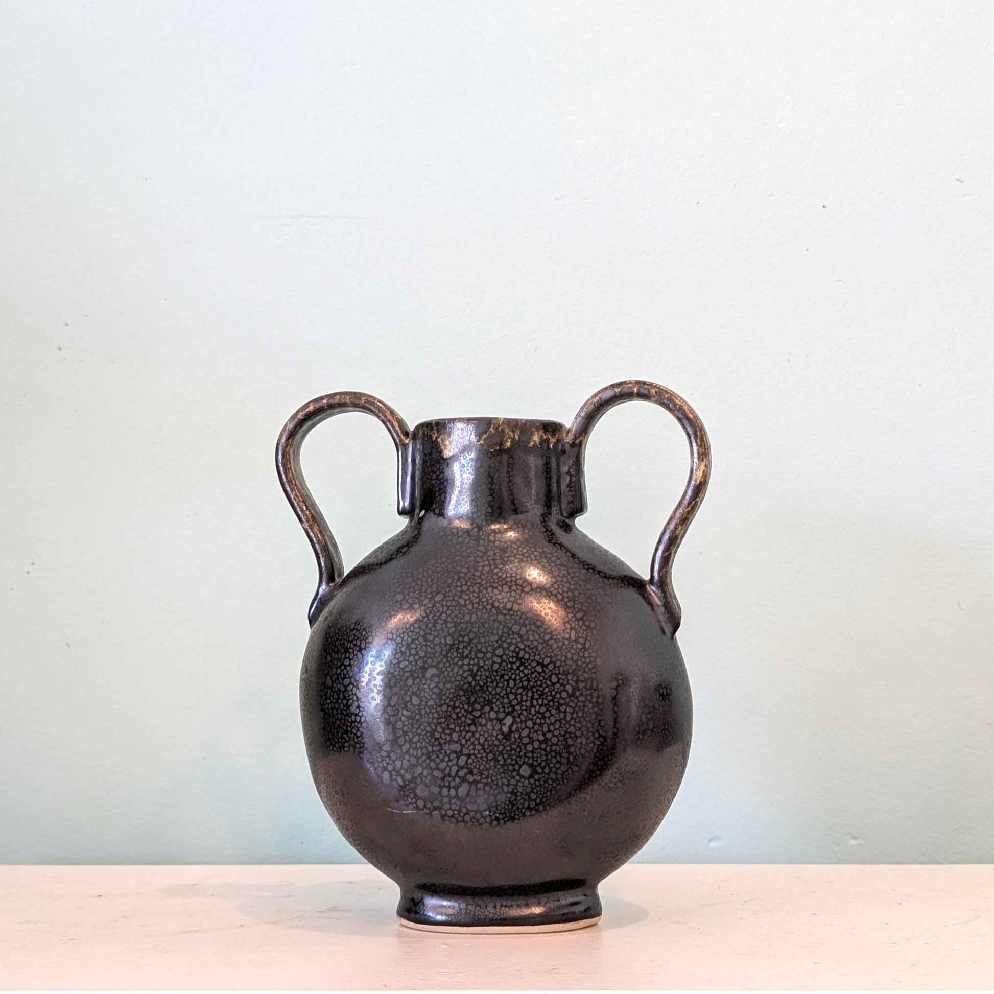 A dark glazed ceramic vase with a rounded body, narrow neck, and two loop handles, shown on a light surface against a pale blue wall.
