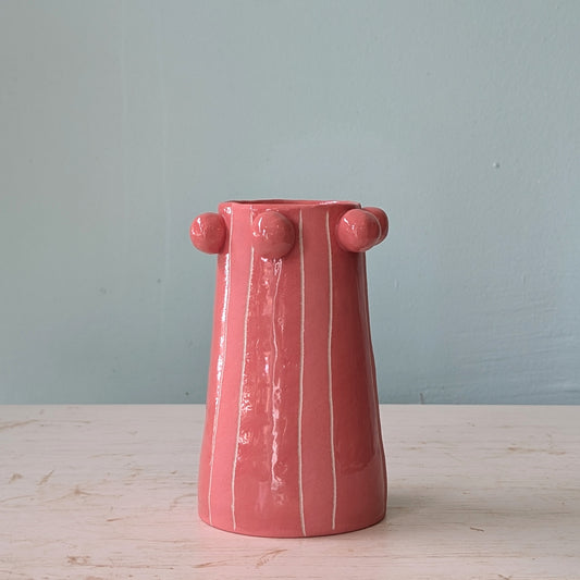 Pink ceramic vase with round bobbles at the top, on a white table against a blue wall.