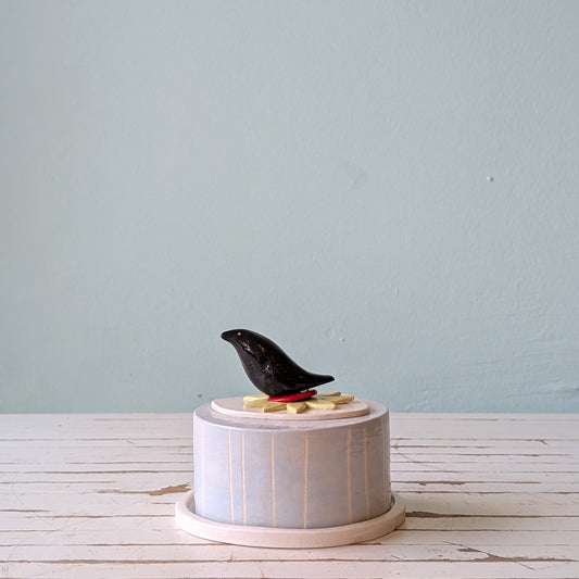 Ceramic butter dish with a black bird handle on a white pedestal against a light blue wall.