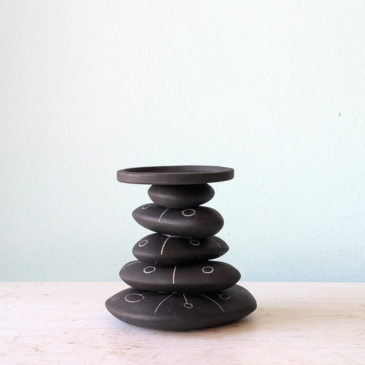 A black ceramic stack candlestick with white hand-drawn line details, displayed on a light-colored wooden surface against a pale blue wall.