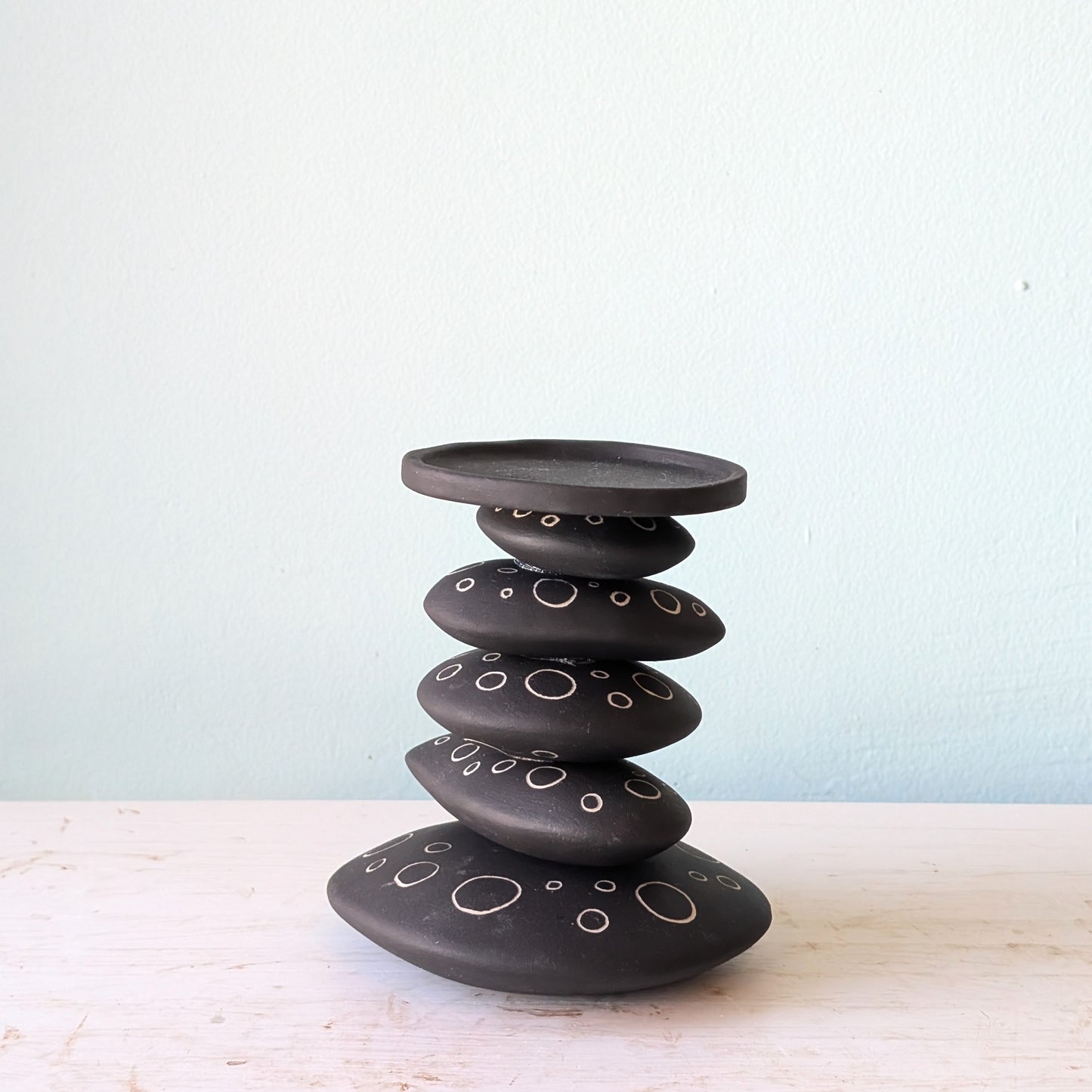 A black ceramic stack candlestick with white hand-drawn circle details, displayed on a light-colored wooden surface against a pale blue wall.