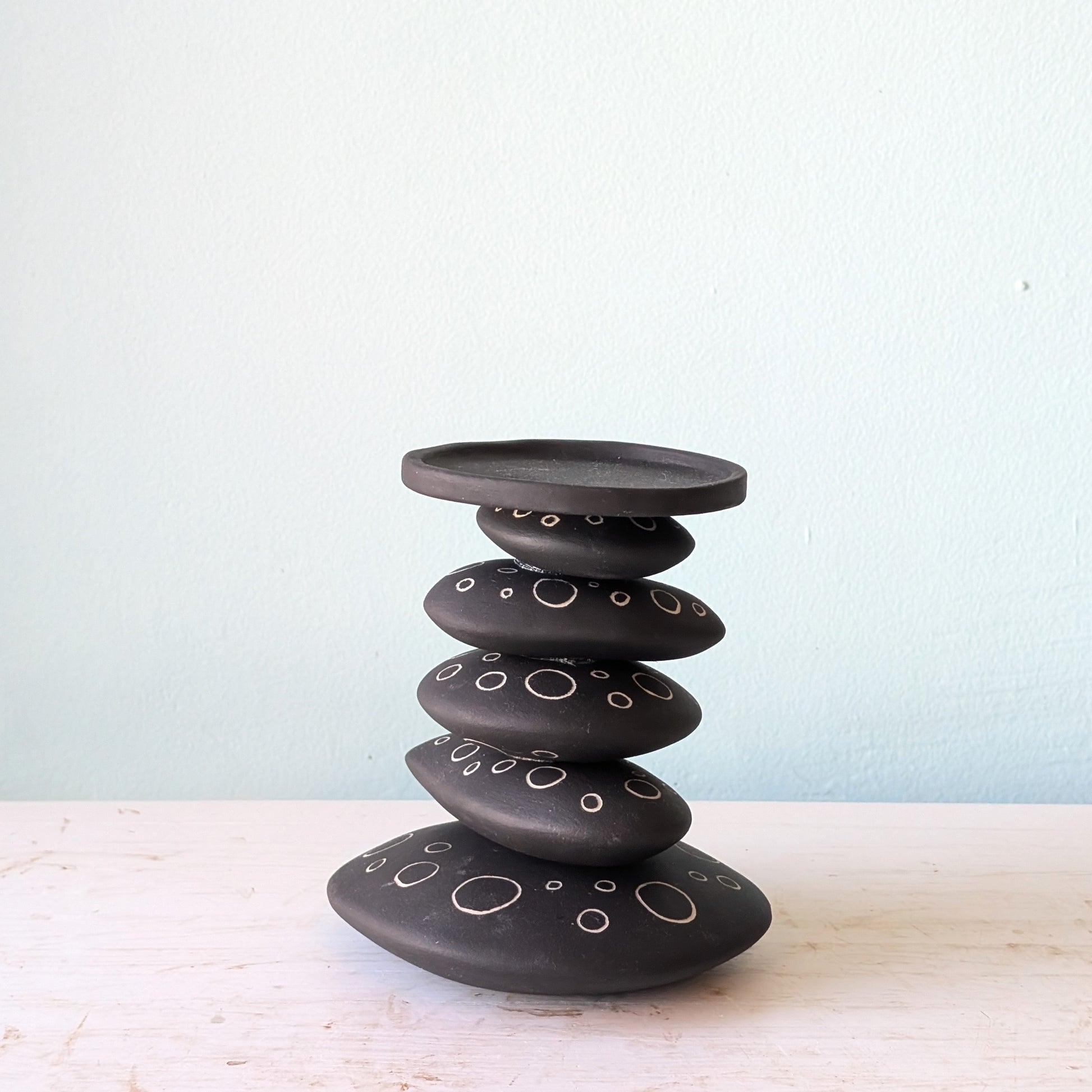 A black ceramic stack candlestick with white hand-drawn circle details, displayed on a light-colored wooden surface against a pale blue wall.