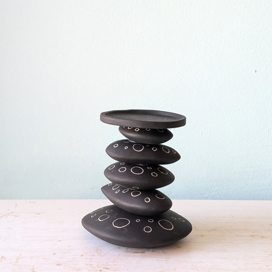 A black ceramic stack candlestick with white hand-drawn circle details, displayed on a light-colored wooden surface against a pale blue wall.