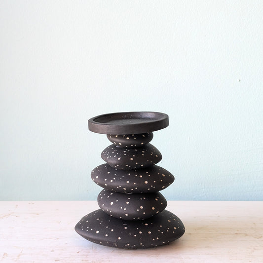 A black ceramic stack candlestick with white hand-drawn dot details, displayed on a light-colored wooden surface against a pale blue wall.
