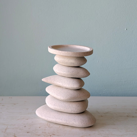 A candlestick in that shape of a stack of organic-shaped ceramic stones in speckled cream-beige, on a light wooden surface against a blue wall.