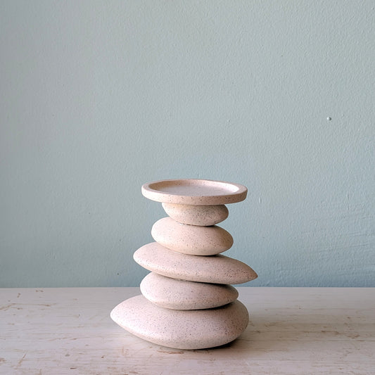 A candlestick in that shape of a stack of organic-shaped ceramic stones in speckled cream-beige, on a light wooden surface against a blue wall.