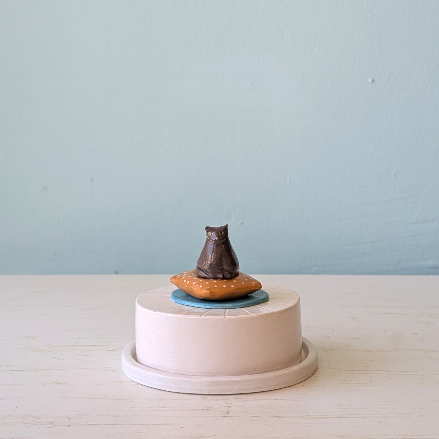 Small round butter dish with a cat figurine on top against a light blue background