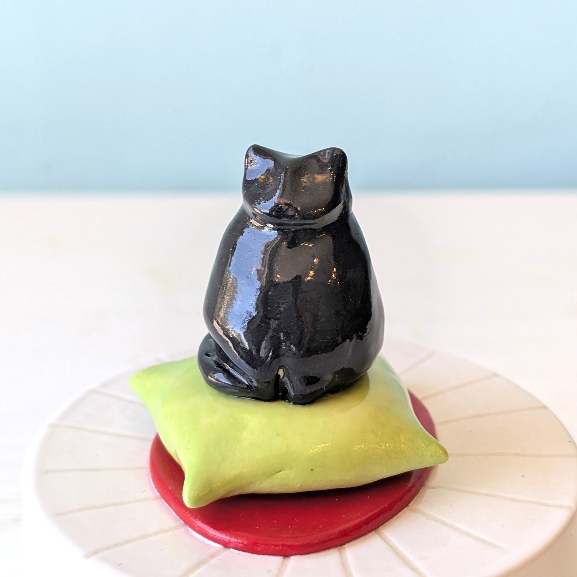 Black cat figurine on a green pillow with a red base against a light blue background