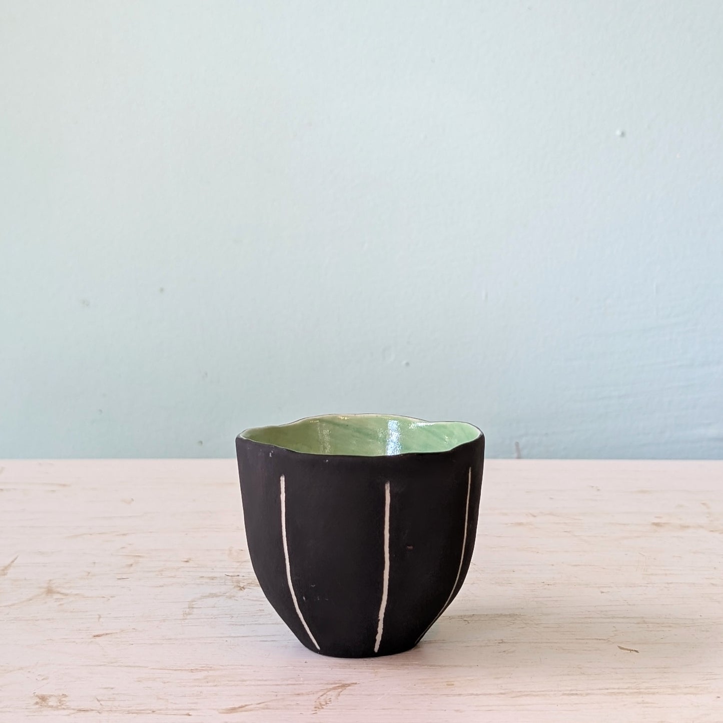 Black ceramic cup with green interior on a light wooden surface against a light blue wall.