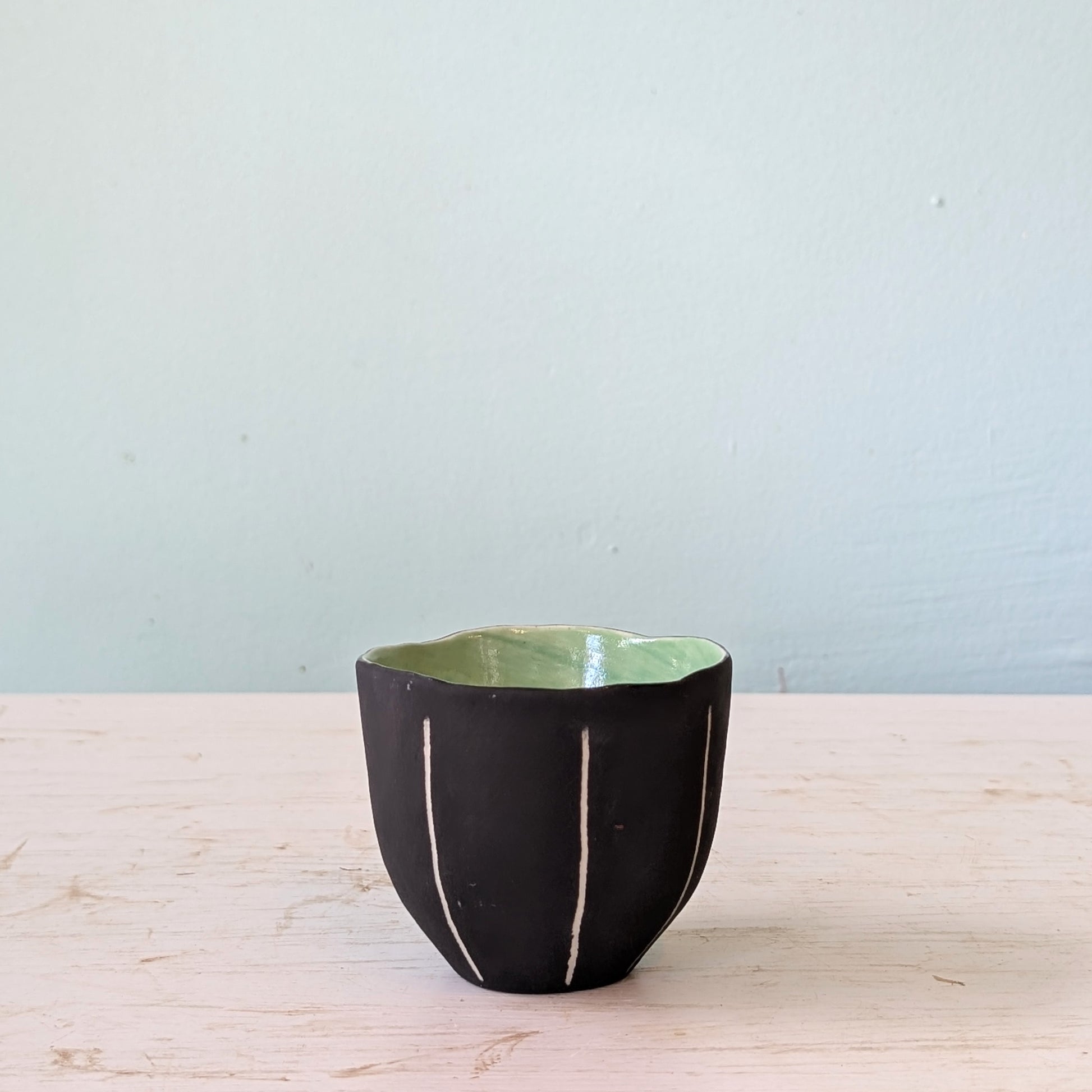 Black ceramic cup with green interior on a light wooden surface against a light blue wall.