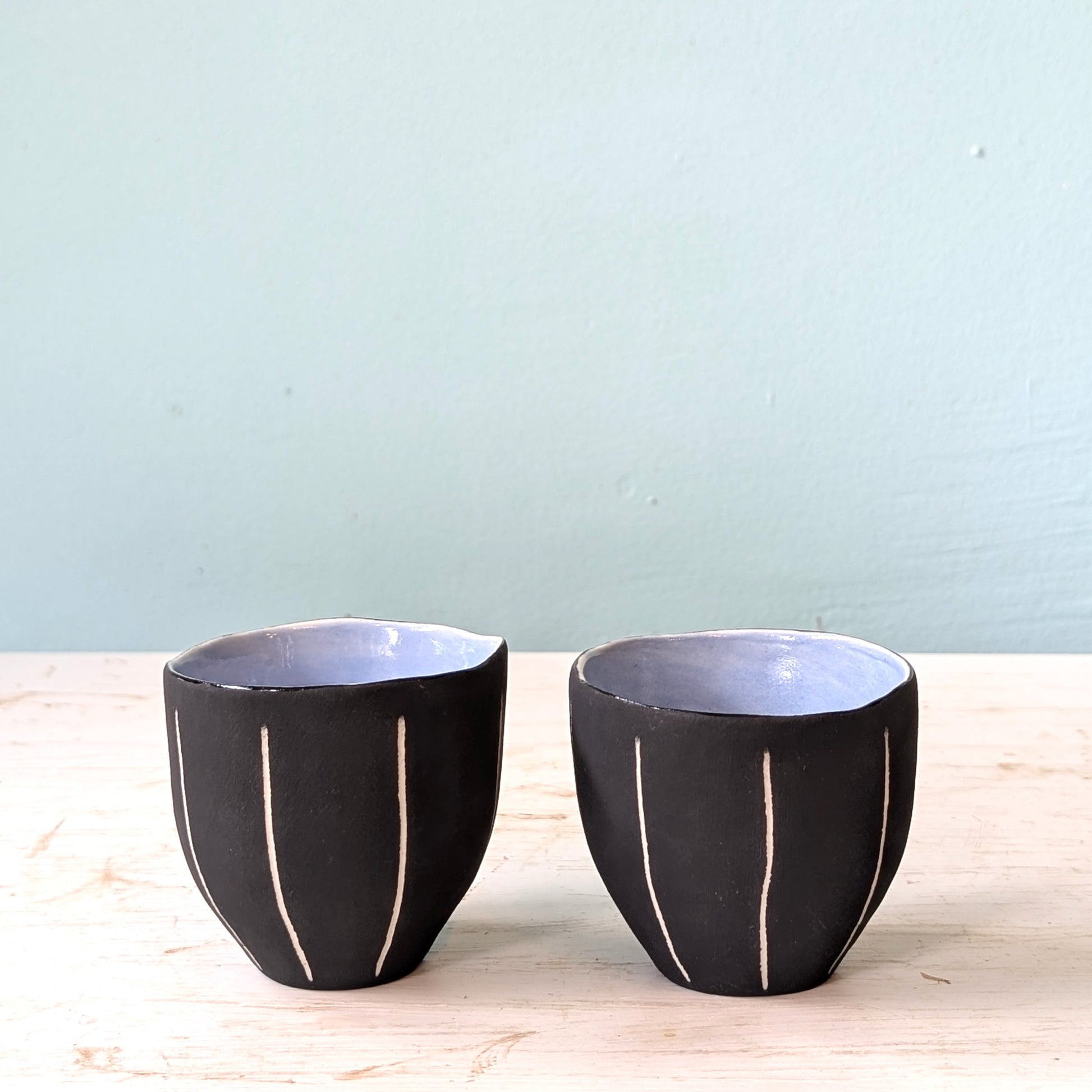 Two black ceramic cups with blue interior on a light wooden surface and light blue background
