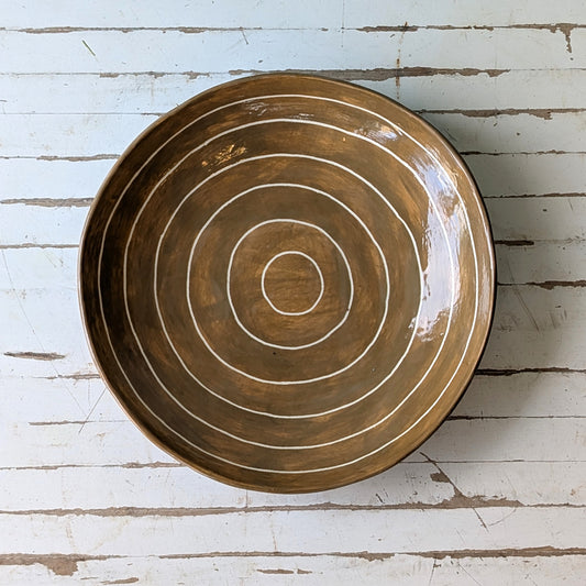 Top view of a brown ceramic plate with carved concentric circles on a white wooden surface.