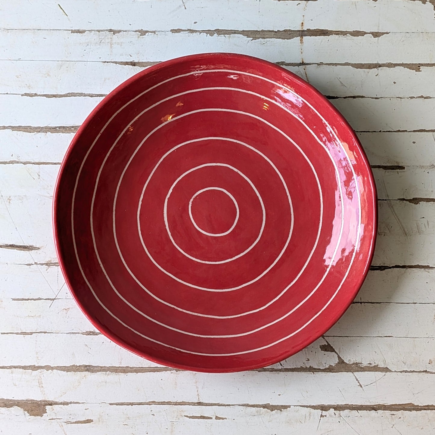 Top view of a red ceramic plate with carved concentric circles on a white wooden surface.