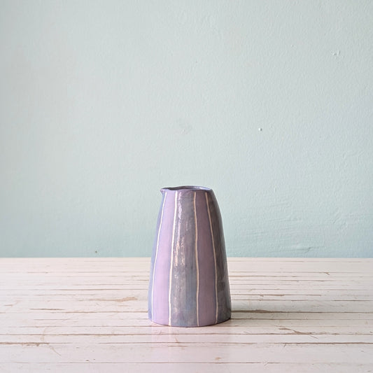 Front view of a lilac ceramic pourer on a white wooden surface against a blue background.
