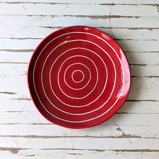 Top view of a red ceramic plate with carved concentric circles on a white wooden surface.