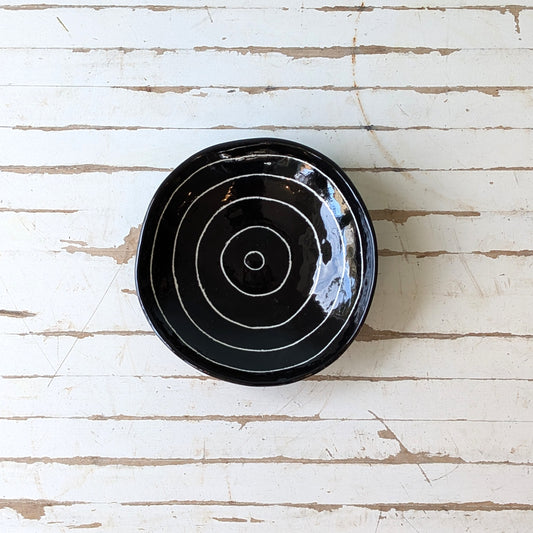 Top view of a black ceramic bowl with carved concentric circles on a white wooden surface.