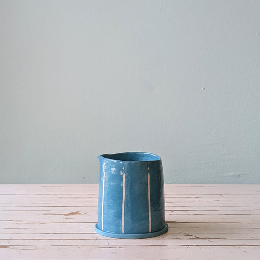 Front view of a blue ceramic pourer on a white wooden surface against a blue background.
