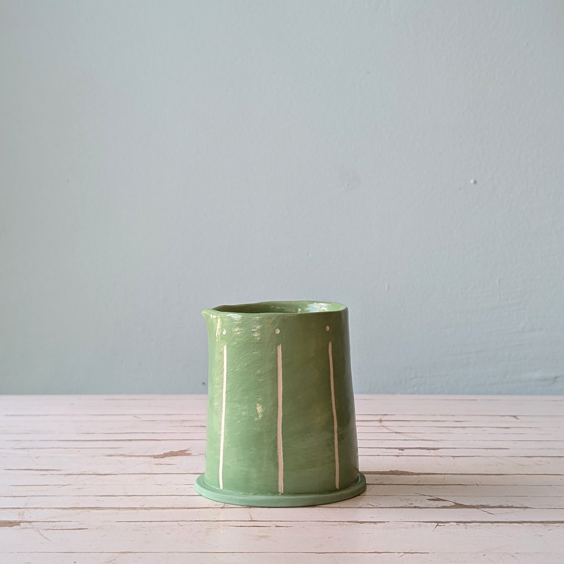 Front view of a green ceramic pourer on a white wooden surface with a blue background.