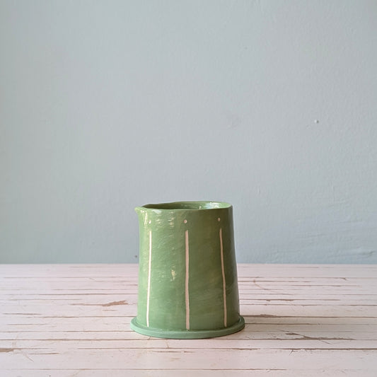 Front view of a green ceramic pourer on a white wooden surface with a blue background.