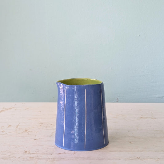 Blue ceramic pourer with green interior on a light wooden surface against a light blue wall.