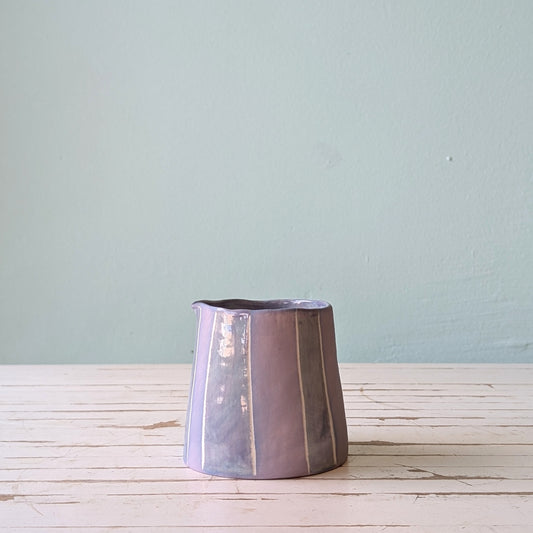 Front view of a lilac ceramic pourer on a white wooden surface against a blue background.